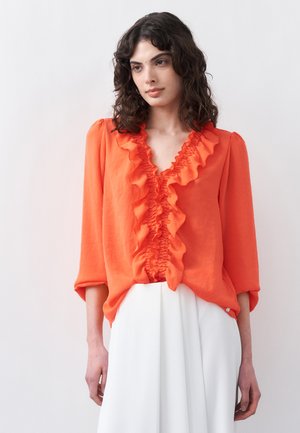 Γυναίκα με σγουρά σκούρα μαλλιά, φορώντας φωτεινή πορτοκαλί μπλούζα με βολάν και λευκό παντελόνι ψηλόμεσο, στέκεται μπροστά σε απλό λευκό φόντο.