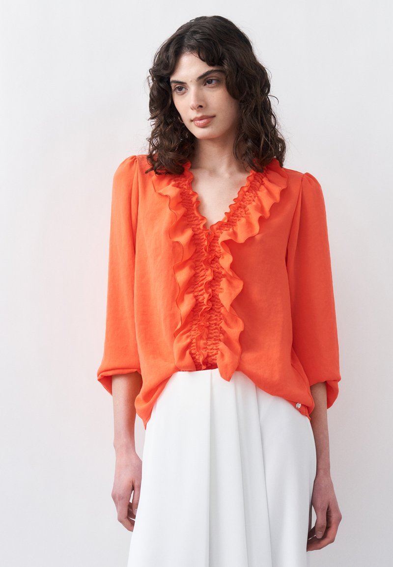 Γυναίκα με σγουρά σκούρα μαλλιά, φορώντας φωτεινή πορτοκαλί μπλούζα με βολάν και λευκό παντελόνι ψηλόμεσο, στέκεται μπροστά σε απλό λευκό φόντο.