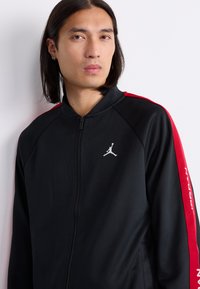 Man with shoulder-length hair wears black zip-up jacket with red stripes on sleeves and white Jumpman logo on chest.
