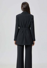 Femme avec de longs cheveux noirs portant un blazer noir ceinturé et un pantalon évasé assorti, se tenant de dos contre un fond clair.