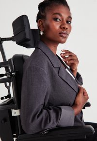 Σκούρο γκρι σακάκι με εφηβικές πέτες, μακρ�ιά μανίκια και λεπτή υφή. Μοντέλο καθισμένο σε αμαξίδιο δύναμης, προβάλλοντας τη στάση των χεριών και τις μοναδικές λεπτομέρειες σχεδίασης.