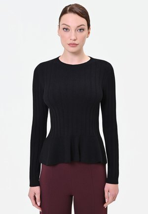 Donna con capelli tirati all’indietro che indossa un maglione nero a coste aderente con orlo a peplo e pantaloni rosso scuro, in piedi su sfondo bianco.