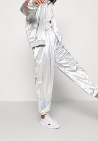 Chándal plateado brillante con puños elásticos; presenta acentos gráficos en azul y rojo en los pantalones. Combinado con zapatillas deportivas blancas.