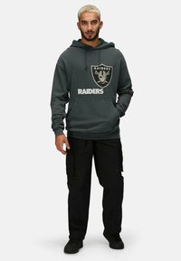 Sudadera gris con el logo de los Raiders, bolsillo frontal y cordón ajustable. Combinada con pantalones cargo negros y deportivas negras.