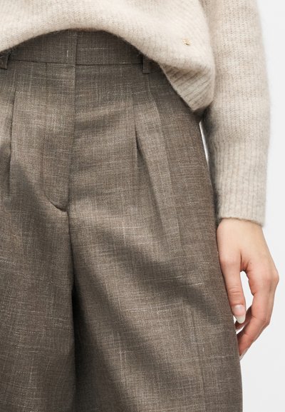 Μπεζ πουλόβερ σφιχτά μέσα σε ψηλόμεση καφέ υφασμάτινη φαρδιά παντελόνα, με εμφανές χέρι να ακουμπάει δίπλα στο ισχίο.