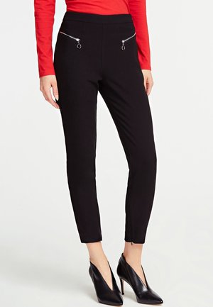 Mujer con pantalones negros ajustados con bolsillos de cremallera plateada, top rojo de manga larga y zapatos negros de tacón alto puntiagudos sobre fondo liso.