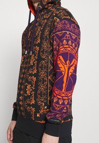 Felpa con cappuccio con base nera, intricate fantasie floreali arancioni, dettagli viola sulle maniche, con un design centrale di figura.