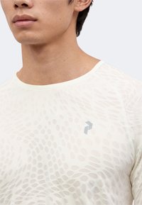 Gros plan d'une personne portant une chemise beige clair à motifs avec un petit logo réfléchissant sur la poitrine, sur un fond uni.