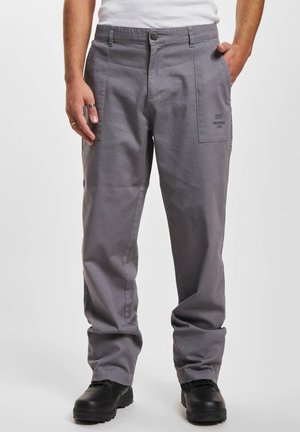 Pantalons en coton gris avec une coupe ample, dotés de deux poches latérales, d'une fermeture à bouton et d'un détail logo près de la poche. Des chaussures noires complètent la tenue.