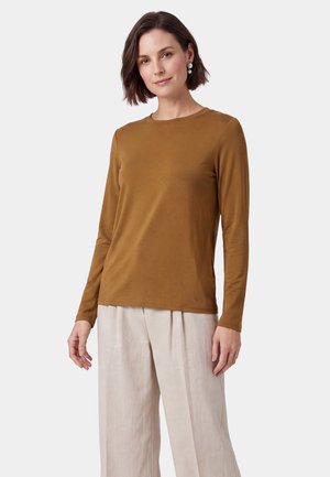 Frau mit kurzen braunen Haaren, die Perlenohrringe, ein braunes Langarmshirt und hellbeige, weit geschnittene Hosen vor einem schlichten Hintergrund trägt.