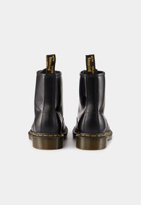 Bottines en cuir noir avec une finition lisse, des coutures jaunes et une semelle texturée. Comprend des tirettes noires avec du texte jaune.