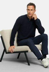 Marineblaues Langarm-Henley-Shirt mit einer Drei-Knopf-Leiste, kombiniert mit marineblauen Hosen und weißen Sneakers, sitzt auf einem modernen hellbeigen Stuhl.