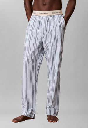 Osoba bez koszulki, ubrana w białe spodnie do spania w pionowe czarne paski z kremowym pasem Calvin Klein, stojąca z rękami w kieszeniach.