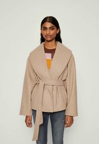 Μπεζ περιτυλιγμένο παλτό με φαρδιές πέτες και ζώνη, με μαλακή υφή. Φοριέται πάνω από ένα πολύχρωμο πουλόβερ με σχέδια, σε συνδυασμό με τζιν.