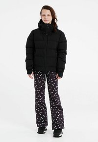 Schwarze Daunenjacke mit hohem Kragen und Reißverschluss. Kombiniert mit schwarzen Hosen mit einem weißen Blumenmuster. Schwarze Stiefel runden den Look ab.