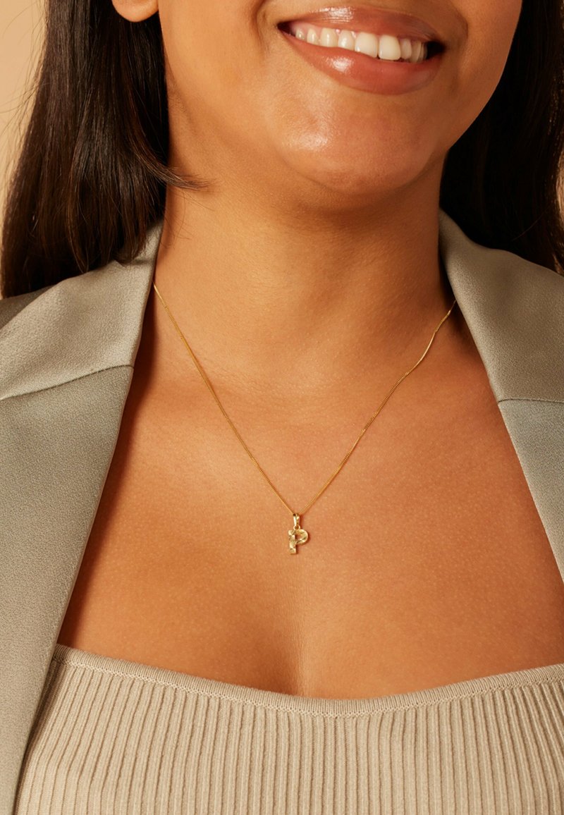 Gouden ketting met een gebogen hanger met een klein bedeltje, gedragen over een geribbelde top in neutrale kleur en onder een lichtgrijze blazer.
