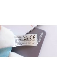 White plush toy with a soft blue patterned fabric, featuring a label with care instructions and certifications, placed on a dark background.