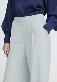 Šviesiai pilki aukštai užtraukiami kelnių su šoninėmis kišenėmis, raukšlėtu priekiniu dizainu ir lygiu paviršiumi, derinami su tamsiai mėlynu satino bluzonu.