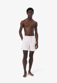 Costume da bagno rosa chiaro con cintura elastica e cordoncino, caratterizzato da un piccolo logo verde. Il modello è in piedi, scalzo e posato in posizione eretta.