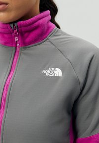 Primer plano de una persona que lleva una chaqueta de forro polar gris y morada con cremallera frontal y el logo de The North Face en el pecho.