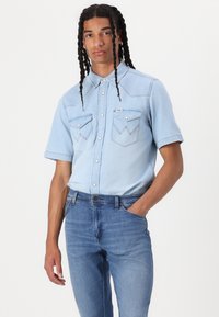 Camicia di jeans azzurro chiaro a maniche corte con bottoni a pressione, due tasche sul petto con alette angolari, indossata con jeans di jeans azzurro chiaro.