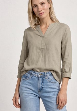 Femme blonde portant un haut beige à manches longues en V et un jean bleu clair, debout devant un fond clair uni.