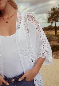 Kimono bianco in pizzo con motivi intricati, tessuto leggero e maniche fluenti, indossato sopra una semplice canotta bianca e pantaloni shorts in denim.