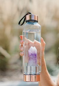 Bouteille d'eau en plastique transparent avec une forme hexagonale, des accents en rose doré et une pierre précieuse violette à l'intérieur. Comprend une sangle de transport noire.