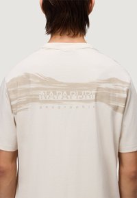 T-shirt in cotone color crema con un motivo astratto beige e il testo "NAPAPIJRI geographic" sul retro in alto. Maniche corte.
