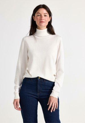 Kvinde med langt mørkt hår iført en hvid højhalset sweater og mørkeblå jeans, stående mod en ensfarvet lys baggrund.