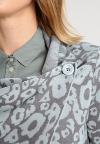 Šedá pletená bunda s světle modrým květinovým vzorem, s výrazným modrým knoflíkem a širokým límcem, navlečená přes zelenou košili.