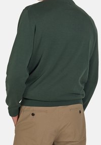 Maglione verde scuro con collo e polsini a coste, abbinato a pantaloni beige chiaro con tasche laterali e dettagli con bottoni.