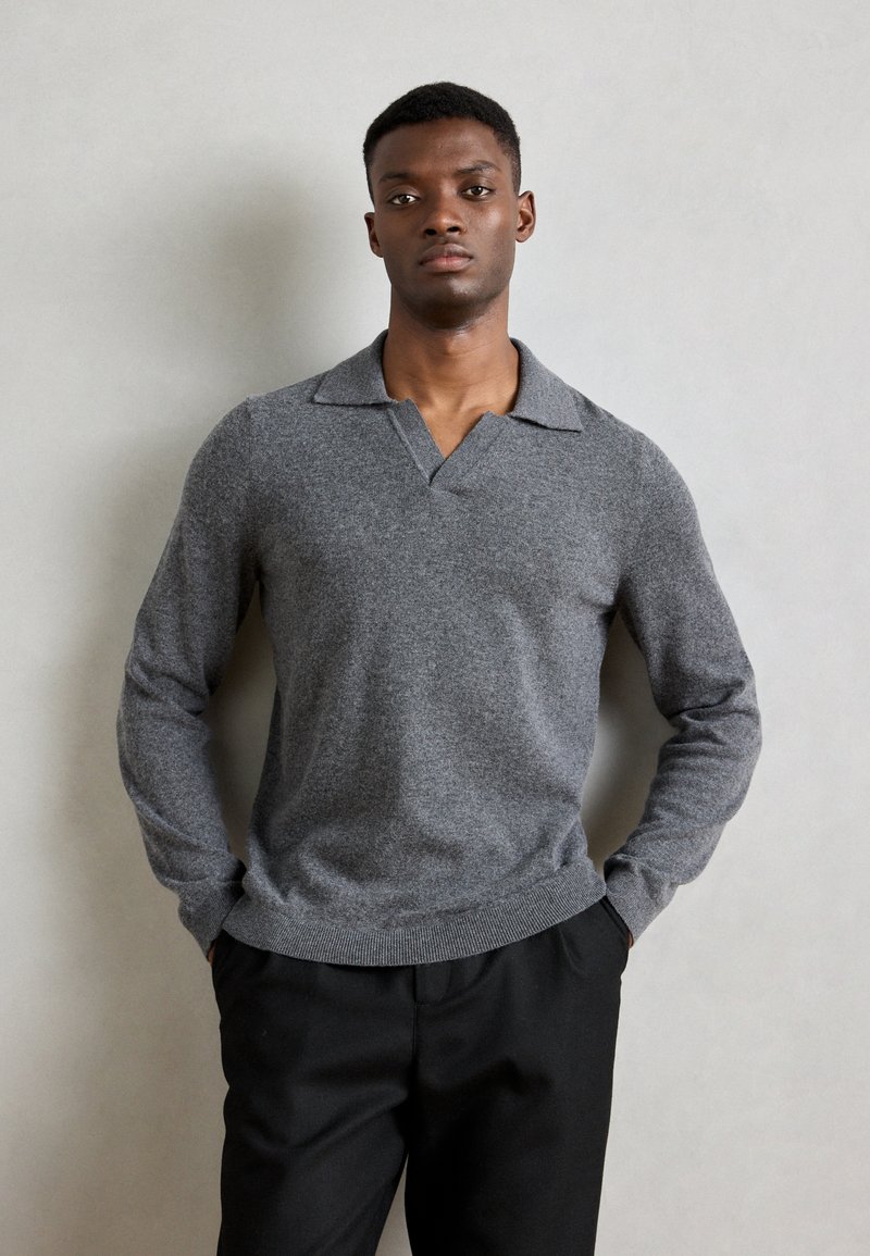 Pull en maille gris à manches longues avec un col et un ourlet côtelé. Présente une texture douce et une coupe décontractée. Un pantalon noir complète le look.