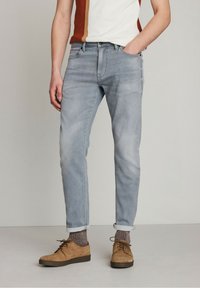 Jeans de mezclilla azul claro con un ajuste relajado, con los puños doblados en los tobillos. Llevados con zapatos de ante marrones y calcetines con estampado.