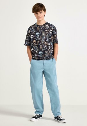 Jonge persoon staand met handen in de zakken, gekleed in een zwart shirt met grafische schedelprint, lichtblauwe broek en zwarte sneakers, tegen een witte achtergrond.