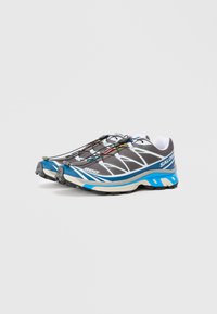 Sapatos de desporto cinzentos com detalhes em azul, apresentando um superior em malha, sola texturizada e atacadores elásticos para um ajuste seguro. Logótipo e padrões de design notáveis.