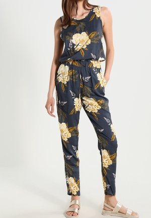 Frau trägt marineblauen ärmellosen Jumpsuit mit großem gelbem Blumenprint und beigefarbenen Sandalen, steht mit einer Hand in der Tasche.