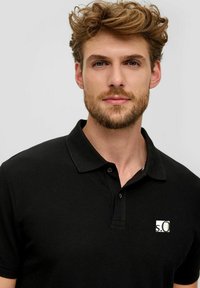 Schwarzes Poloshirt aus strukturiertem Stoff mit einem Dreiknopfverschluss und einem kleinen weißen Logo auf der linken Brust.