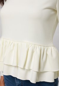 Top blanco de manga larga con un cuerpo ajustado y un dobladillo con volantes en capas. Hecho de un tejido suave con una textura agradable.