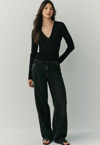 Schwarzes Langarmoberteil mit tiefem V-Ausschnitt, kombiniert mit locker sitzenden, hoch taillierten schwarzen Jeans. Das Model trägt schwarze Sandalen.