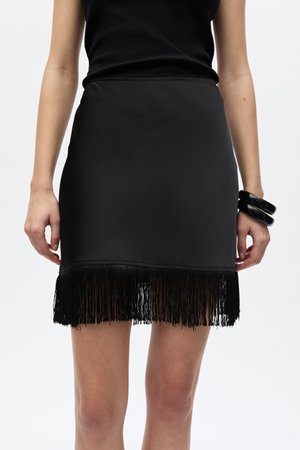 Žena v čiernej mini sukni s strapcovým lemom, čiernom tričku bez rukávov a masívnym čiernym náramkom na pravom zápästí na bielom pozadí.