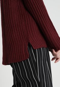 Bordowy sweter o plecionym wzorze z ryżowym splotem, z bocznymi rozcięciami. W zestawieniu z czarno-białymi spodniami w paski. Zbliżenie na rękaw i dół.