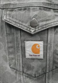 Chaqueta de mezclilla gris con cierre a presión y una etiqueta blanca con un logo naranja y texto rojo. Tejido texturizado con costuras visibles.