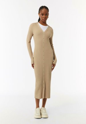 Beige, geripptes Strick-Cardigan-Kleid mit V-Ausschnitt, langen Ärmeln und Knopfleiste, das bis zur Mitte der Wade reicht, kombiniert mit weißen Turnschuhen.