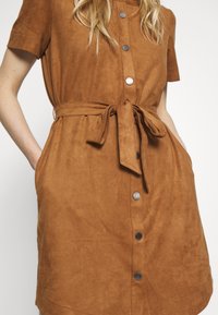 Robe en daim marron avec un col boutonné sur le devant, des manches courtes, une taille ceinturée et des poches latérales. Texture lisse avec une finition douce.
