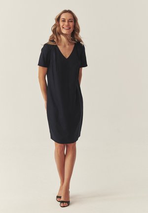 Mujer de pie y sonriendo, vestida con un vestido negro hasta la rodilla con mangas cortas y tacones abiertos negros sobre un fondo liso.