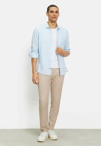 Hellblaues Langarmhemd über einem weißen T-Shirt, kombiniert mit beigen Hosen und weißen Turnschuhen. Glatter Stoff mit Knopfdetails, lässige Passform.