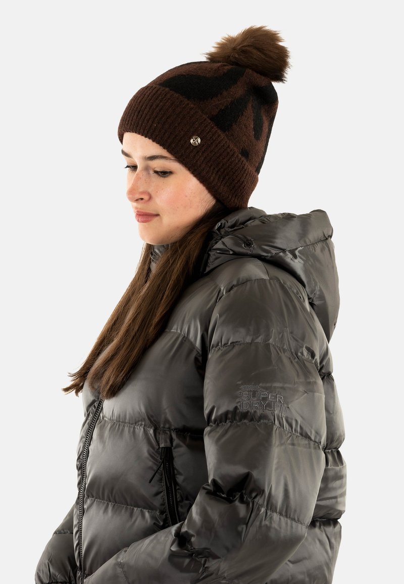 Beanie marrone con motivi neri e un pom-pom sopra, abbinato a una giacca trapuntata lucida di grigio scuro con cappuccio e dettagli con zip.