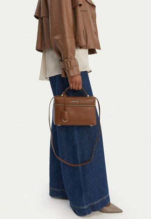 Person wearing brown leather jacket, wide-leg blue jeans, and beige pointed shoes, holding a brown leather handbag with zipper and strap.