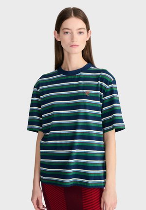 Jeune femme portant un t-shirt rayé bleu marine, vert, blanc et bleu clair avec un petit logo brodé sur la poitrine, debout devant un fond uni.
